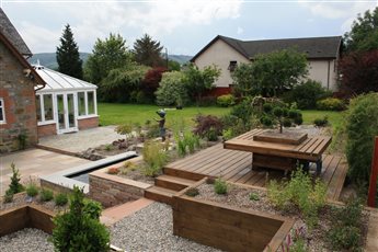 Garden in Fort Augustus. A raised stone pond with a water blade sits between the sandstone terrace and a timber deck, where built-in seating surrounds a cloud-pruned pine, with sleeper beds and gravel planting below.