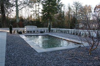 Formal pool to a manor house near Tain. A still, stone-edged rectangle set in gravel reflects mature parkland trees, with clipped evergreens and a simple timber bench giving quiet structure through the winter months.