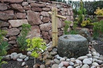 Oriental-inspired garden near Tain. An old granite quern stone, fed by a bamboo spout, forms a quiet water feature against a dry-stone wall, softened by Japanese acers and river cobbles.