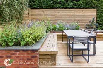 A family garden in Bramhall incorporates a corner seating area with an inbuilt bench over a timber deck. A brick raised planting bed ensures that visitors are surrounded by deliciously scented plants.