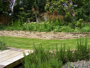 A timber and stone bridge leads across to a lawn, a retaining wall of stone from Ladycross Quarry disappears into the bank. Salvia sclerea 'Turkistanica' stand tall in the borders.