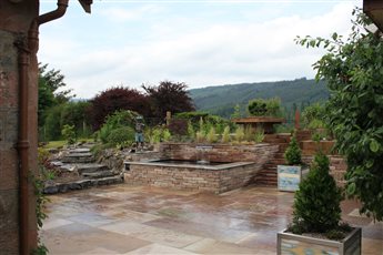 Hillside garden in Fort Augustus with views across forested Highland slopes. A sandstone terrace leads to a raised stone pool with a cascading water blade, framed by stone steps, timber terracing and textured grass planting.
